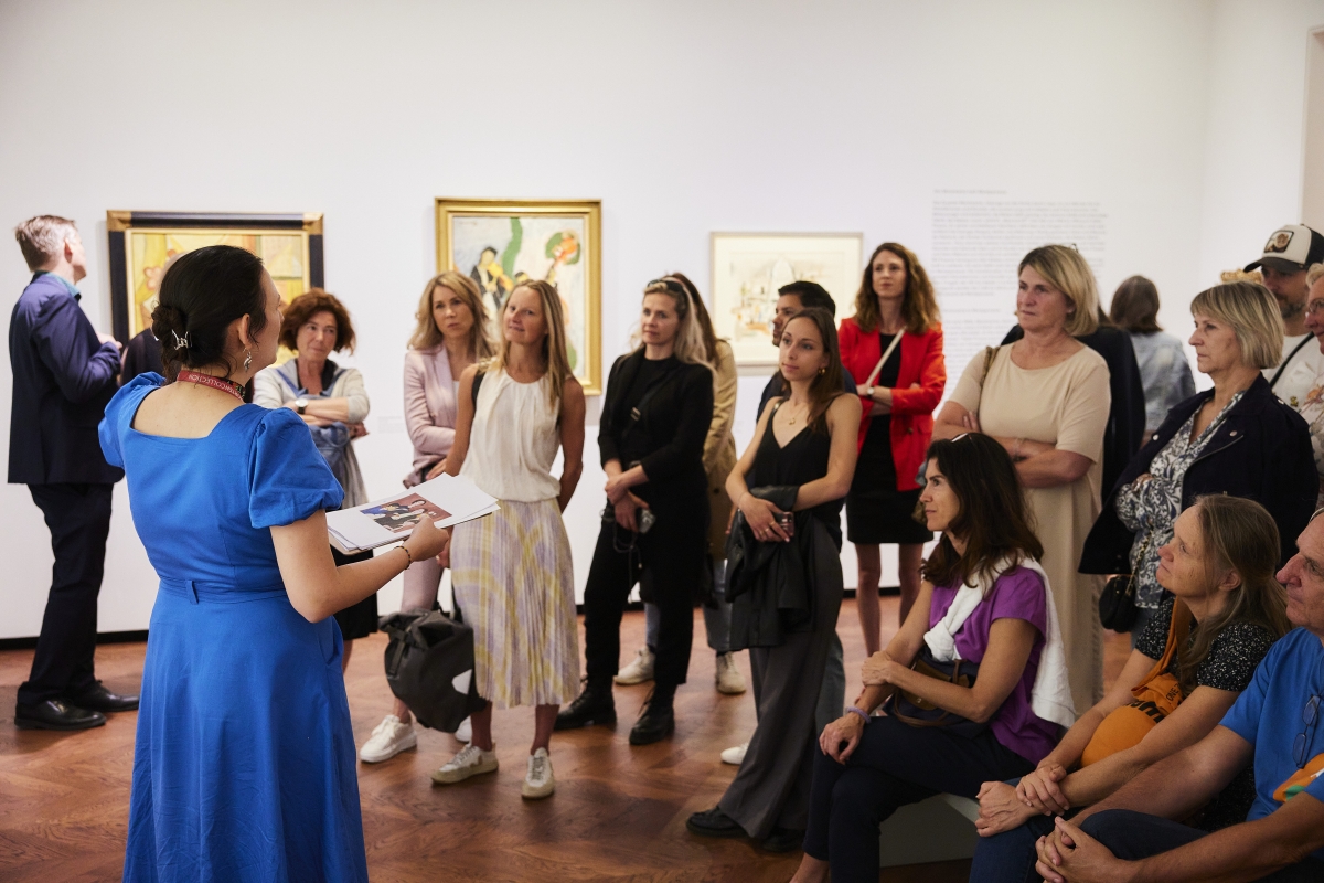 Eine Gruppe von Menschen h&ouml;rt einer Pr&auml;sentation in einer Kunstgalerie zu, umgeben von Bildern an der Wand.