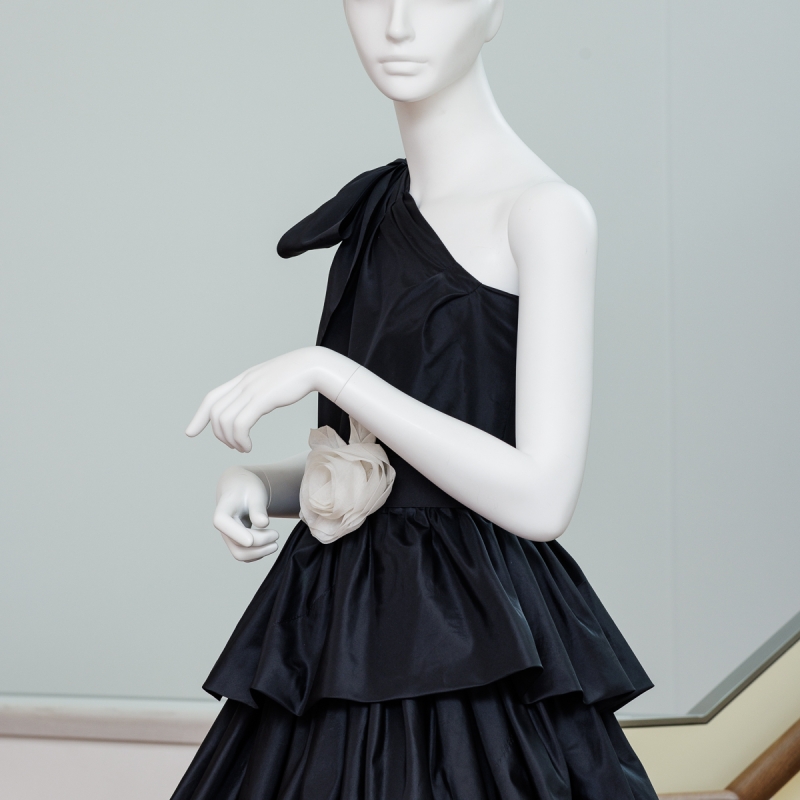 Schwarzes, schulterfreies Abendkleid mit Rüschen und einer blassrosa Blume an der Taille, präsentiert auf einer Schaufensterpuppe.