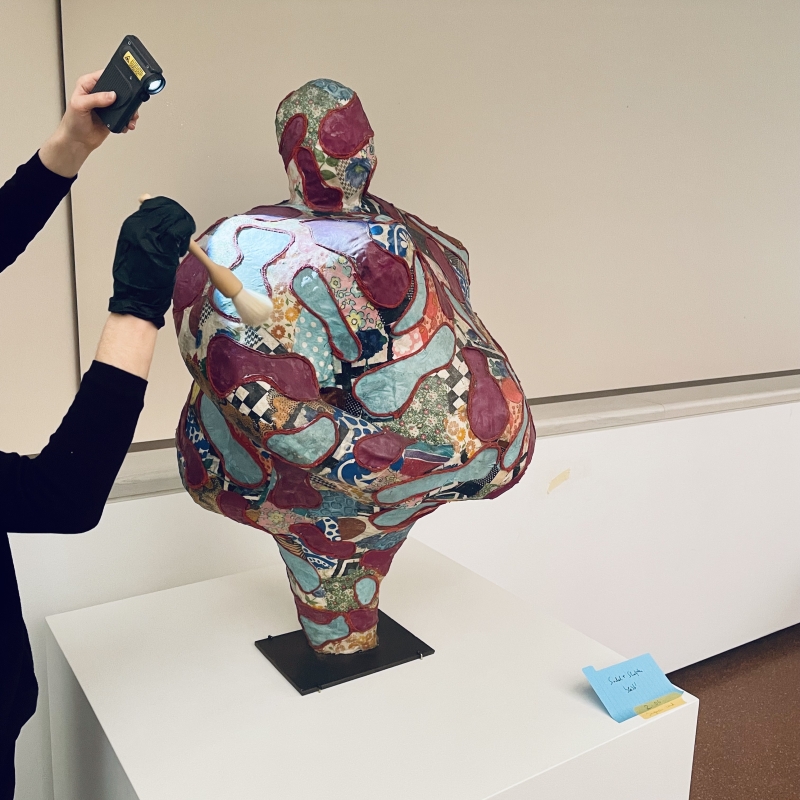 Skulptur eines menschlichen Torso mit bunten, gemusterten Oberfl&auml;chen, w&auml;hrend eine Person sie mit einer Taschenlampe inspiziert.