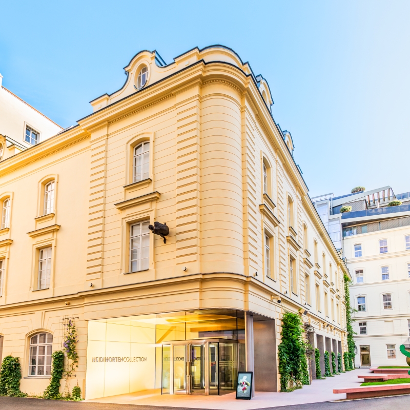 The picture shows the building of the Heidi Horten Collection.
It is located in Hanuschhof, in the center of Vienna.

The building is bright yellow and has a neoclassical façade with large windows.
The shape is reminiscent of an old city palace, but has been renovated in a modern style.

There is a large glass door at the entrance.
Next to it is written in silver lettering: “Heidi Horten Collection”.
A digital screen displays information about the museum.

On the right of the picture you can see a small garden with sculptures.
A large green figure is by the artist Keith Haring.

The photo shows the museum in fine weather.
The sky is blue and the light makes the place friendly and inviting.
