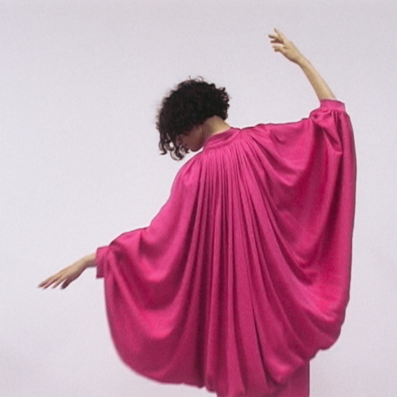 Frau in einem fließenden, pinken Kleid mit weiten Ärmeln, die sich elegant zur Seite dreht.